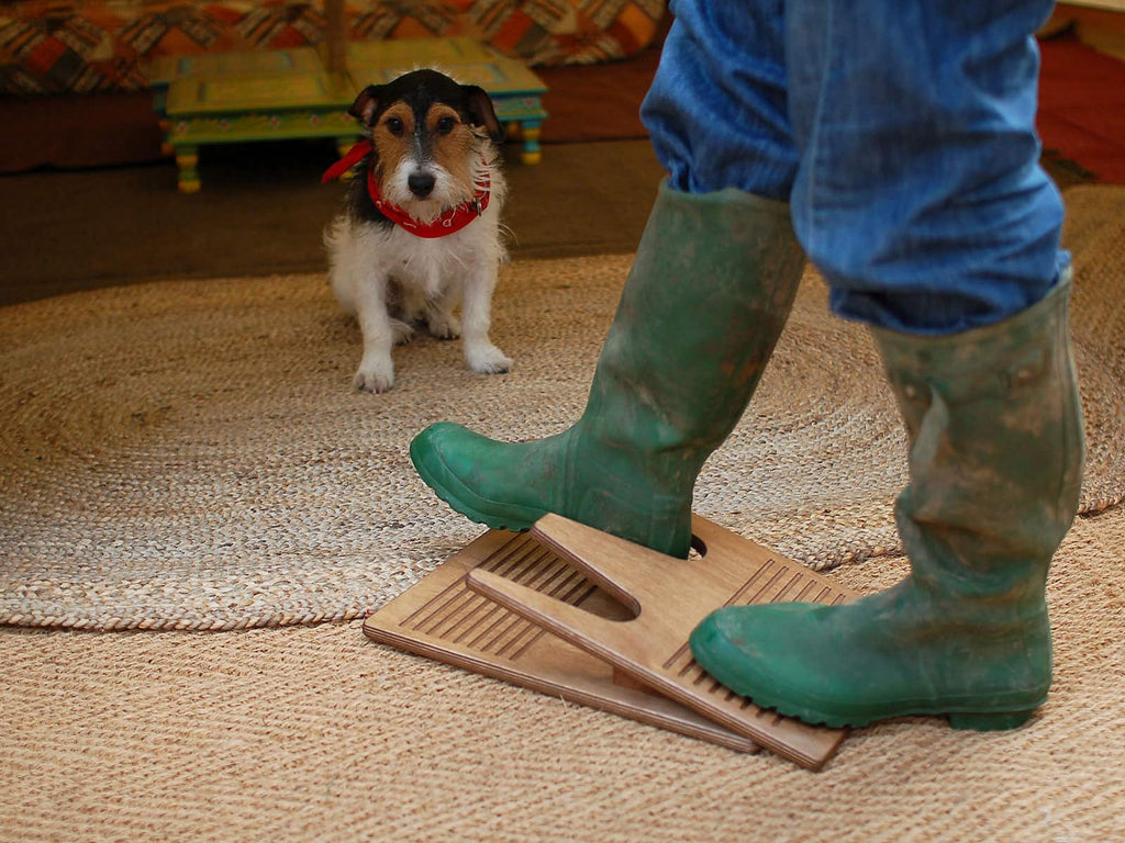 A dog waits patiently sitting on a jute door mat while a person removes their boots